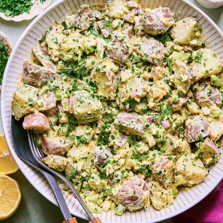 White bowl full of creamy, herby potato salad with serving utensils at the side.