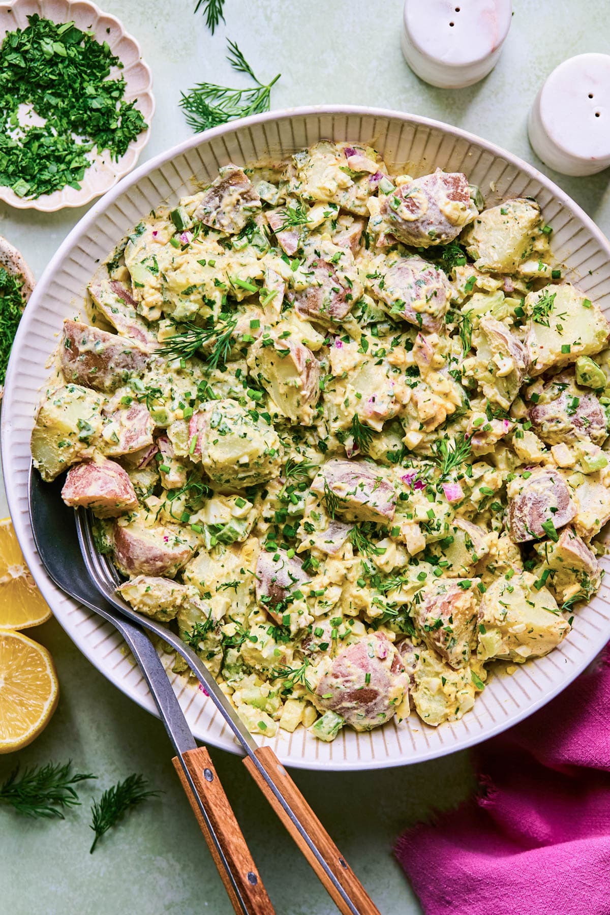 White bowl full of creamy, herby potato salad with serving utensils at the side.