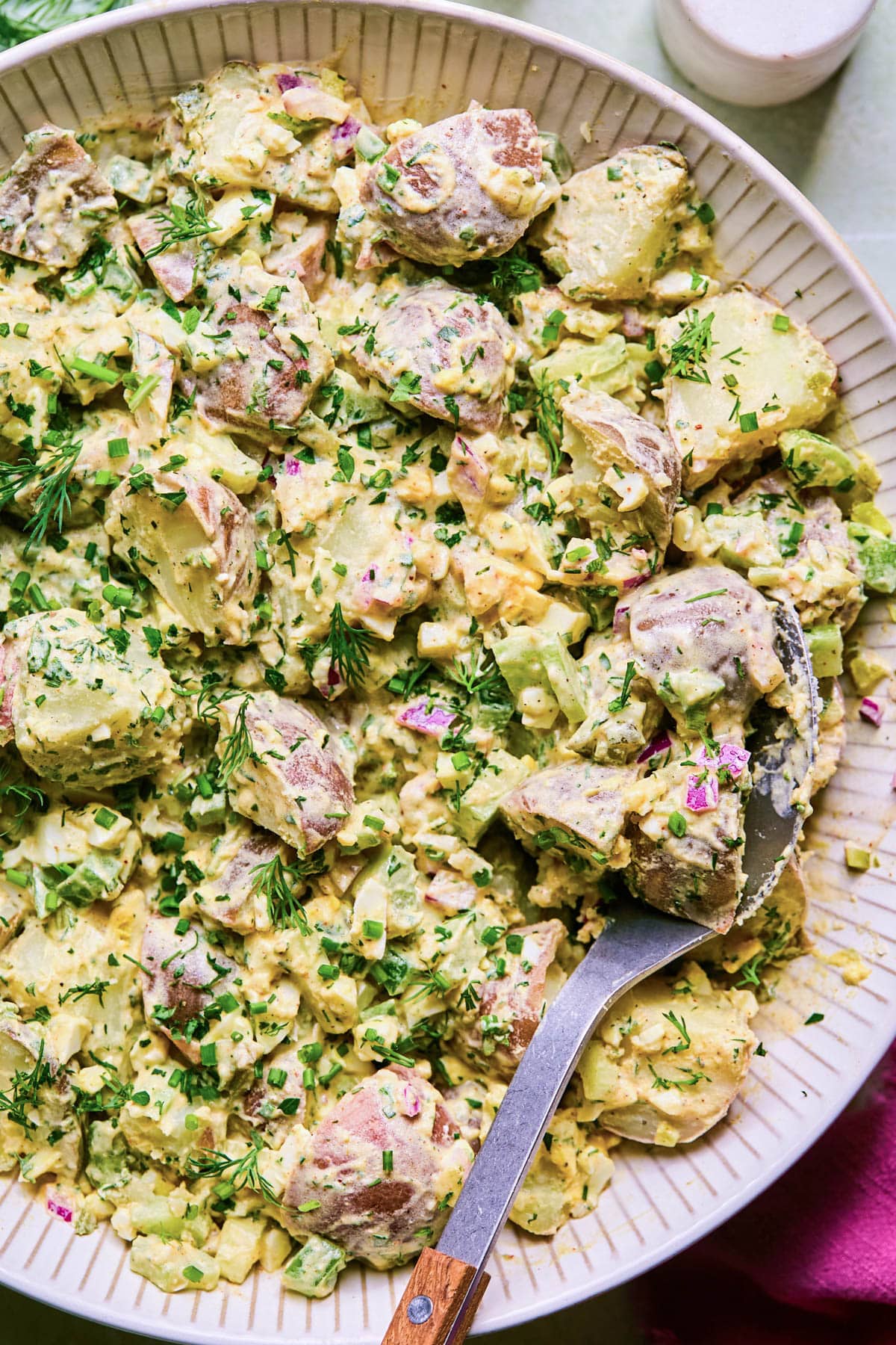 Close up image of a bowl with salad made of chopped red potatoes, creamy dressing, chopped hard boiled eggs, red onion, parsley, chives, pickles, and celery.