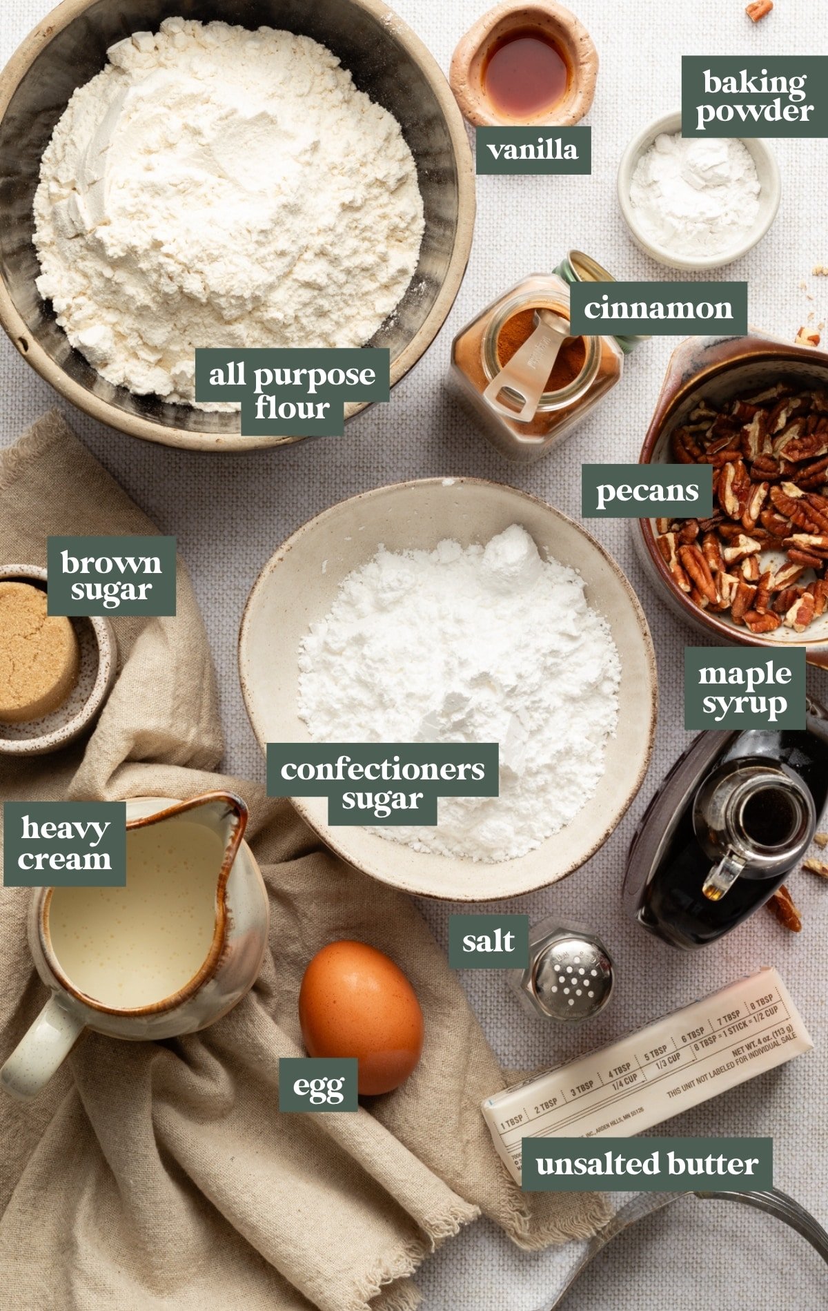 Overhead photo of baking ingredients arranged on a neutral linen background. A large bowl of all-purpose flour sits in the upper left, surrounded by labeled ingredients: vanilla extract, baking powder, ground cinnamon, chopped pecans, maple syrup, brown sugar, confectioners’ sugar, heavy cream, an egg, salt, and a stick of unsalted butter. Each ingredient is neatly styled in ceramic or glass bowls with soft natural lighting, creating a cozy, autumn-inspired baking scene.