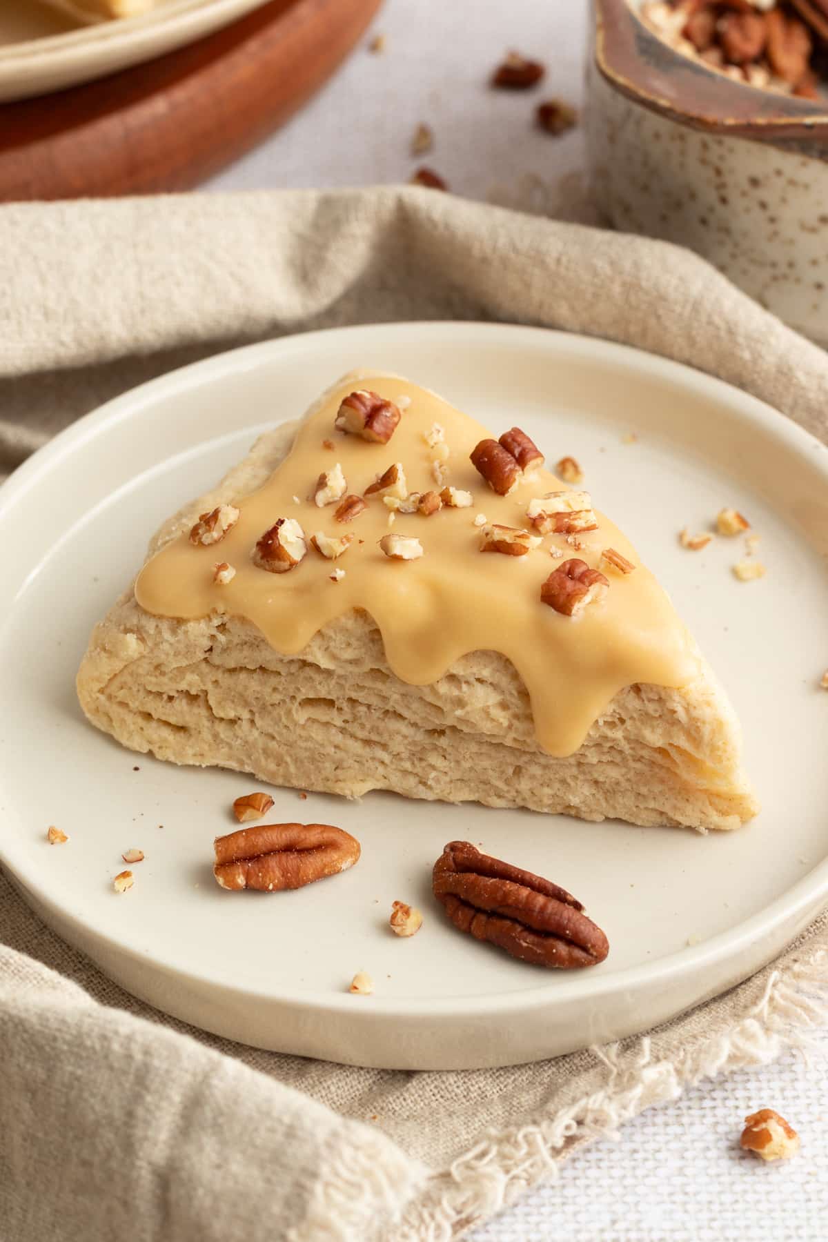A soft maple scone topped with glossy maple glaze and chopped pecans sits on a cream plate with scattered pecan pieces, styled on a neutral linen cloth.