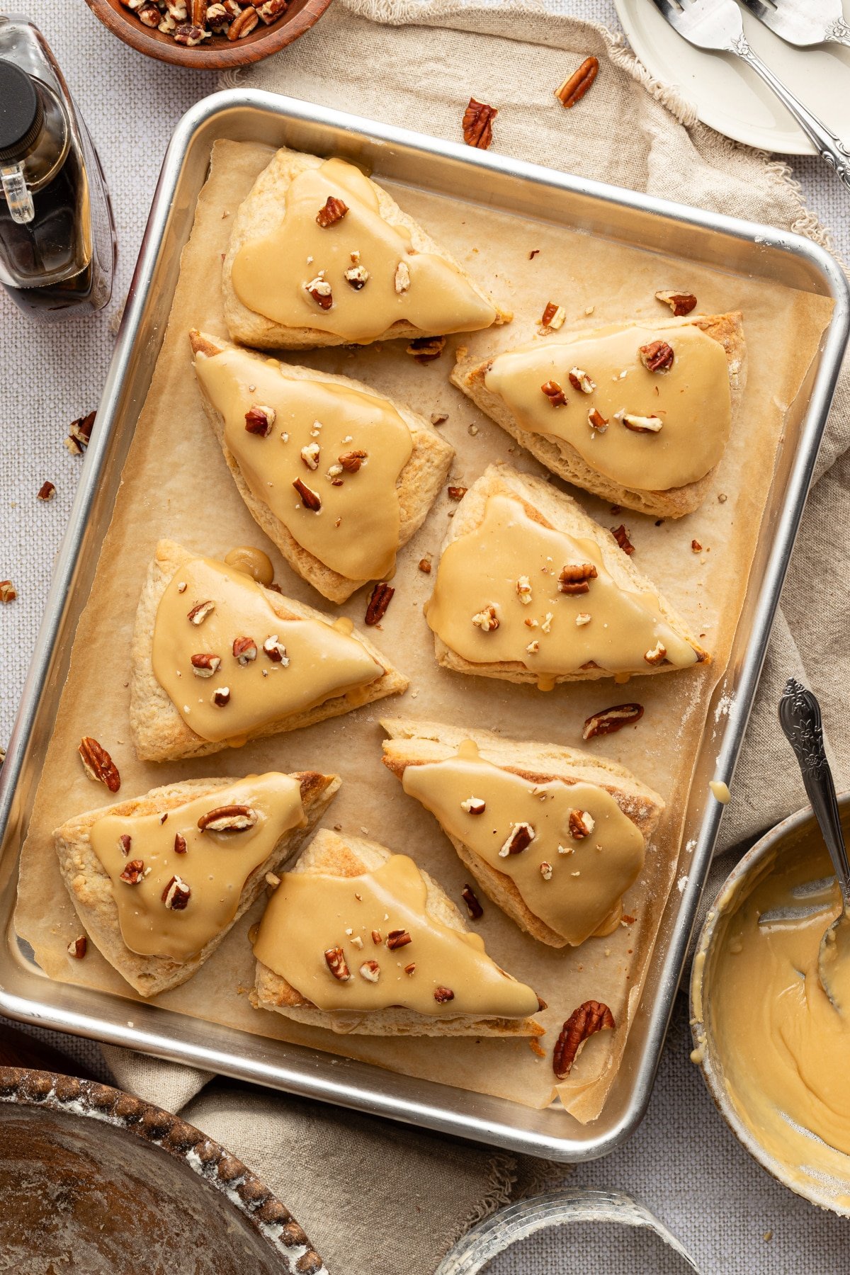 Golden maple scones cooling on a parchment-lined baking sheet, each topped with maple glaze and sprinkled with chopped pecans.