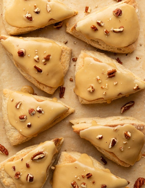 Overhead shot of freshly baked maple scones with smooth maple glaze and pecan pieces on parchment paper, showing their flaky texture.