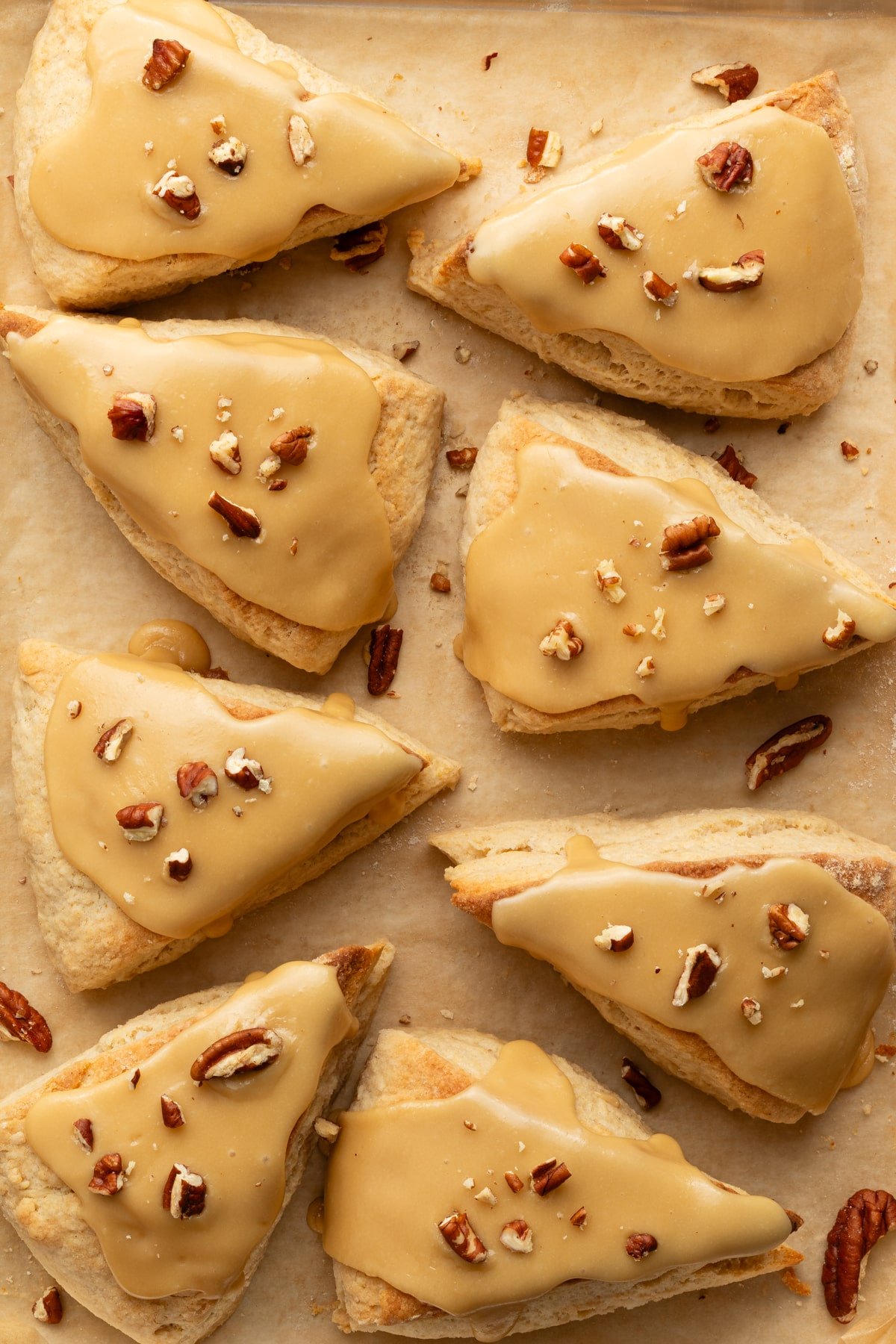 Overhead shot of freshly baked maple scones with smooth maple glaze and pecan pieces on parchment paper, showing their flaky texture.