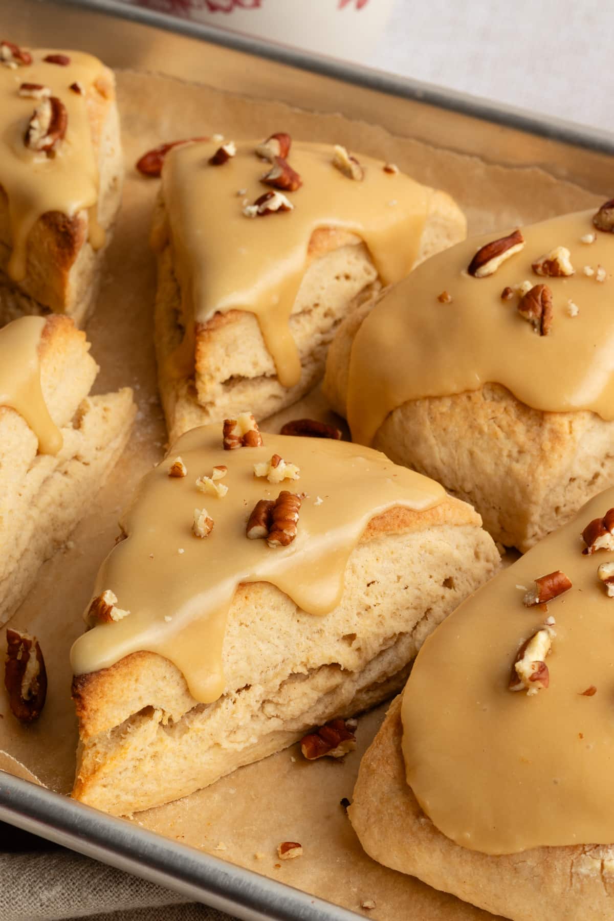 Close-up of thick, tender maple scones on a baking tray, dripping slightly with maple glaze and topped with crunchy pecans.