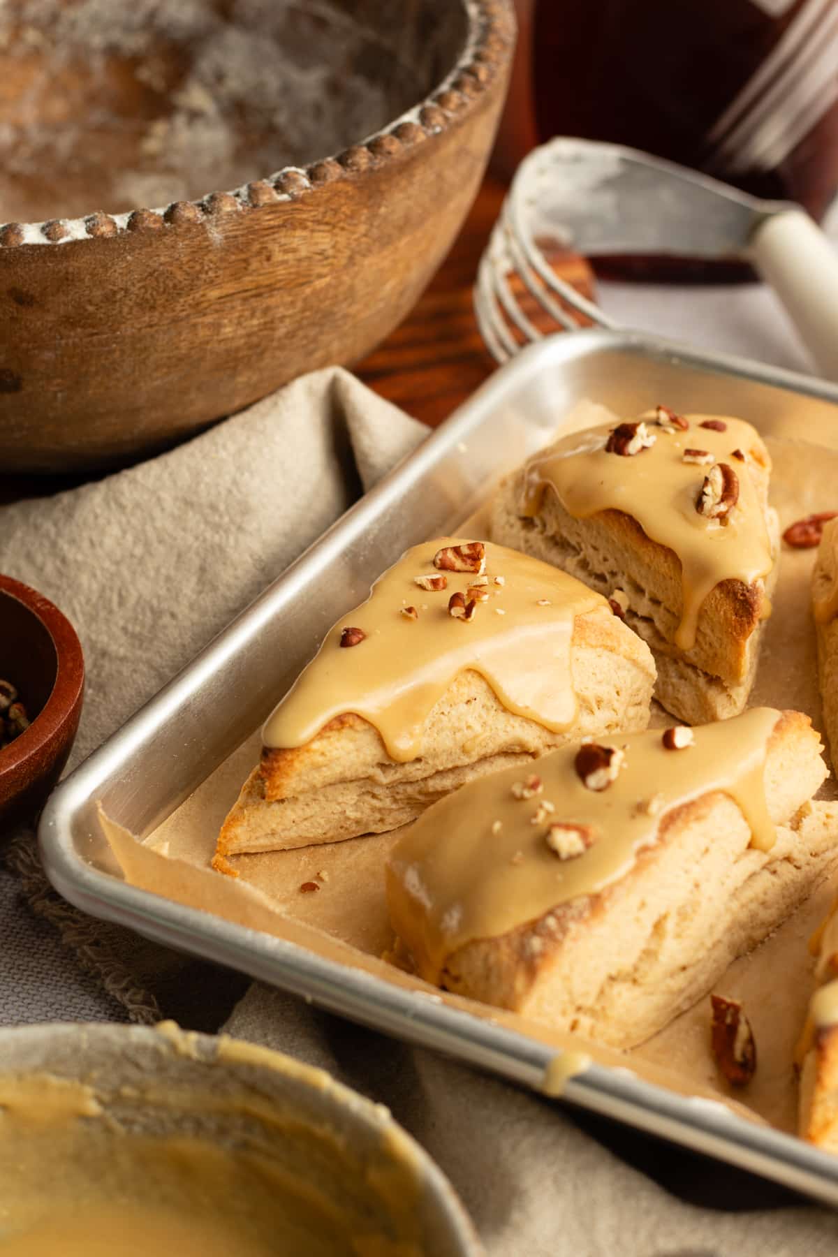 Maple scones on a baking sheet surrounded by a rustic wooden mixing bowl, spoon, and linen — warm, cozy bakery vibes.