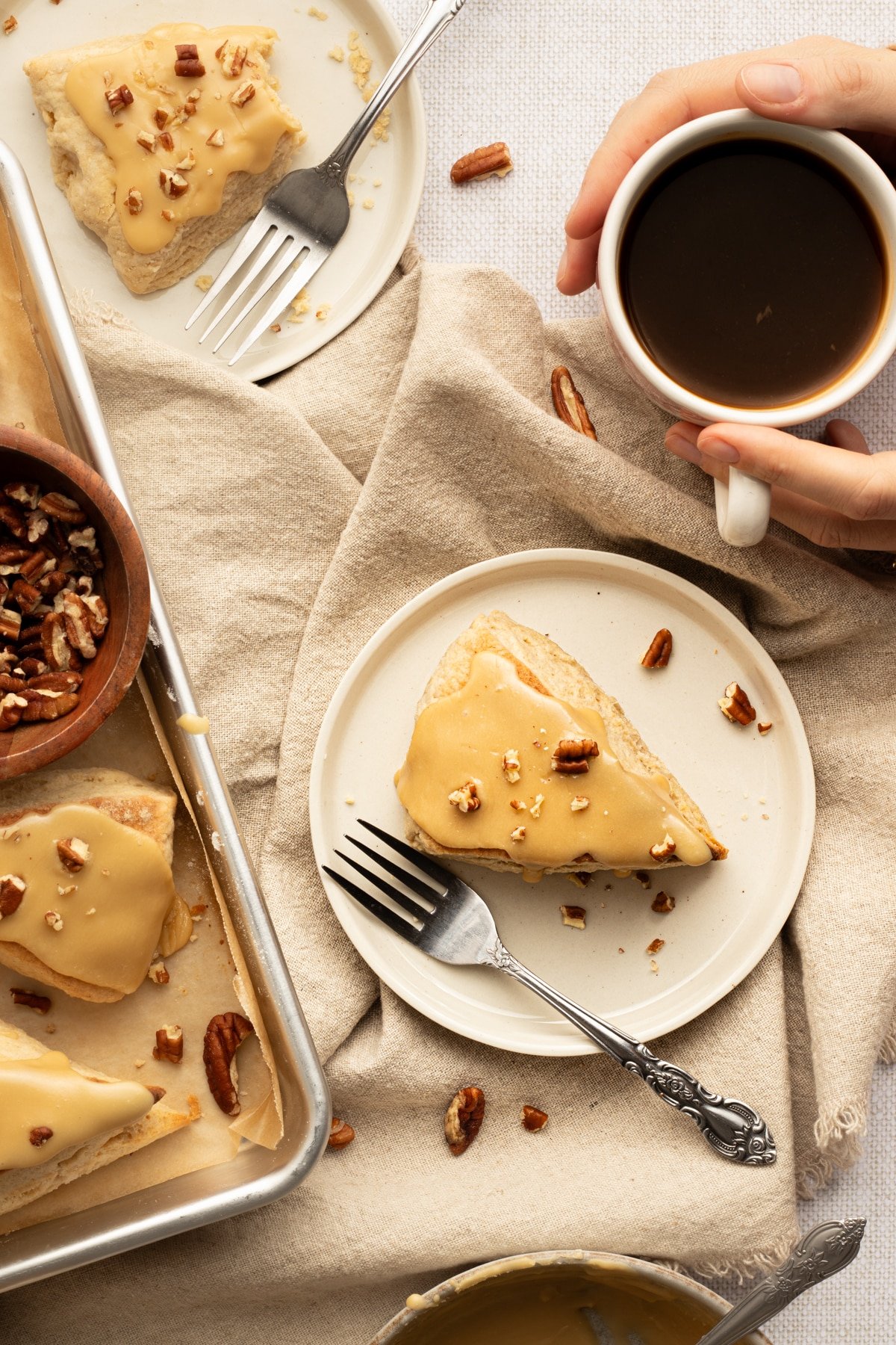 A cozy breakfast scene with glazed maple scones, a mug of black coffee, and scattered pecans on a beige linen backdrop.