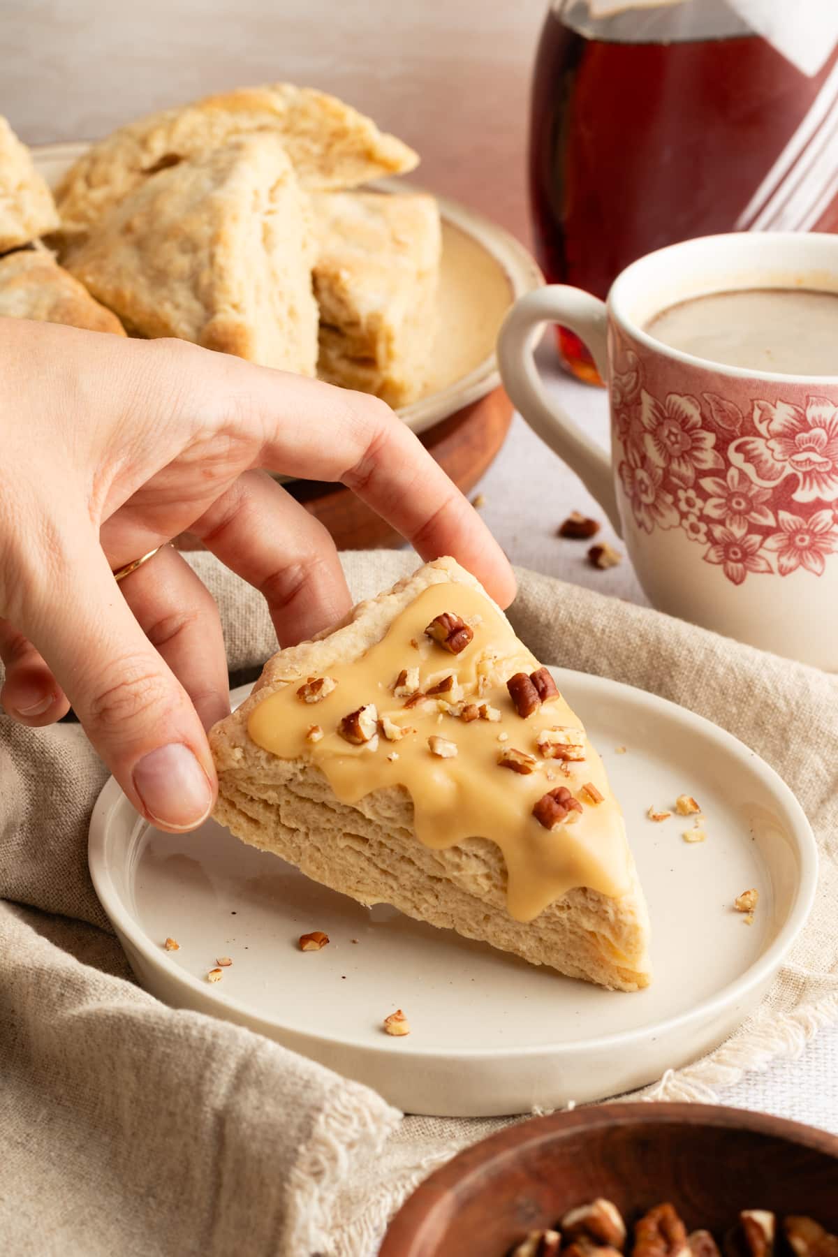 A hand reaching for a maple-glazed scone topped with chopped pecans, with a coffee cup and plate of scones in the background.