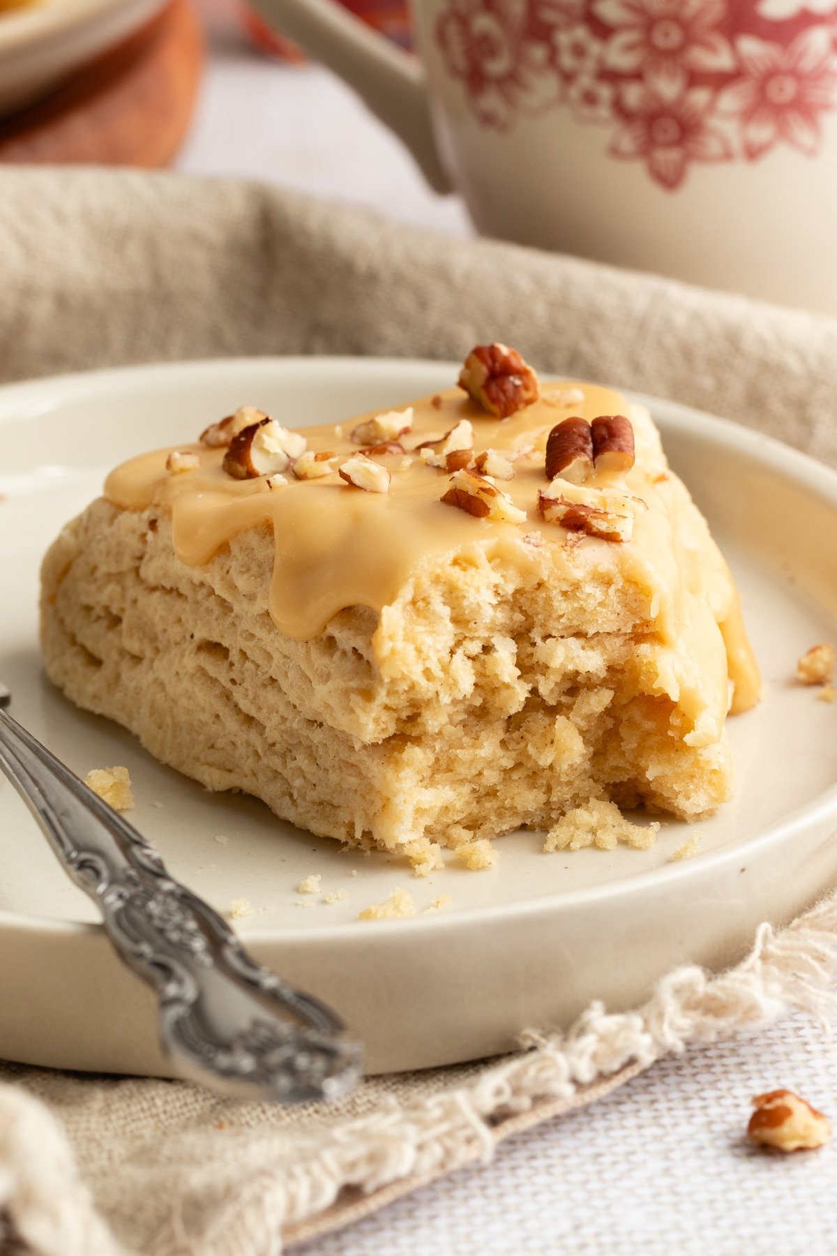 A maple scone with a bite taken out, revealing its tender, fluffy inside, topped with maple glaze and chopped pecans on a plate.