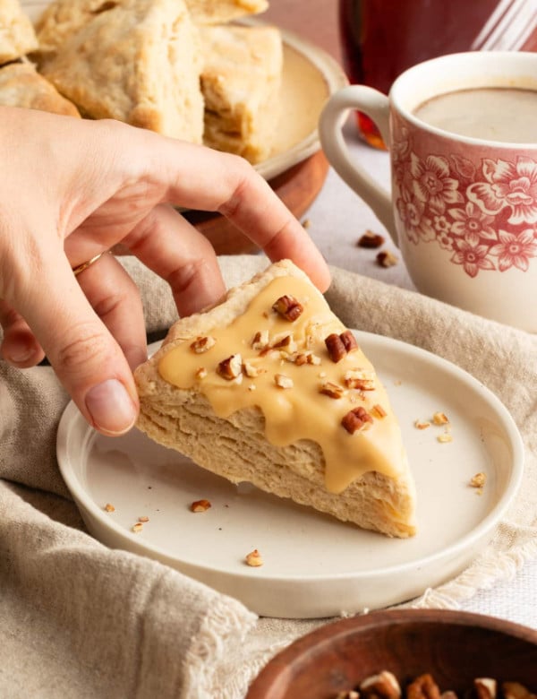 A hand reaching for a maple-glazed scone topped with chopped pecans, with a coffee cup and plate of scones in the background.