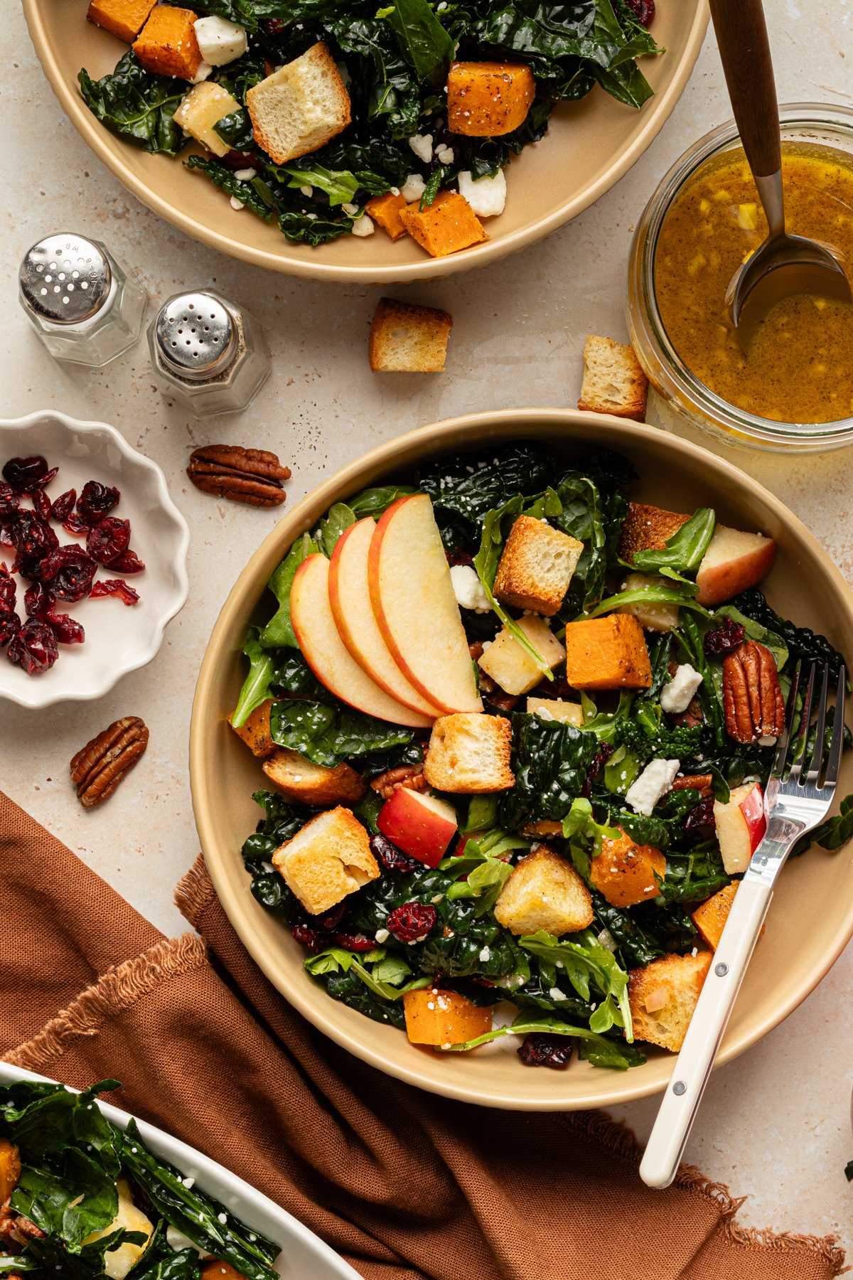 Two bowls of salad filled with kale, roasted squash, apples, pecans, feta, and croutons, surrounded by extra dressing, cranberries, and a brown napkin for a cozy fall presentation.