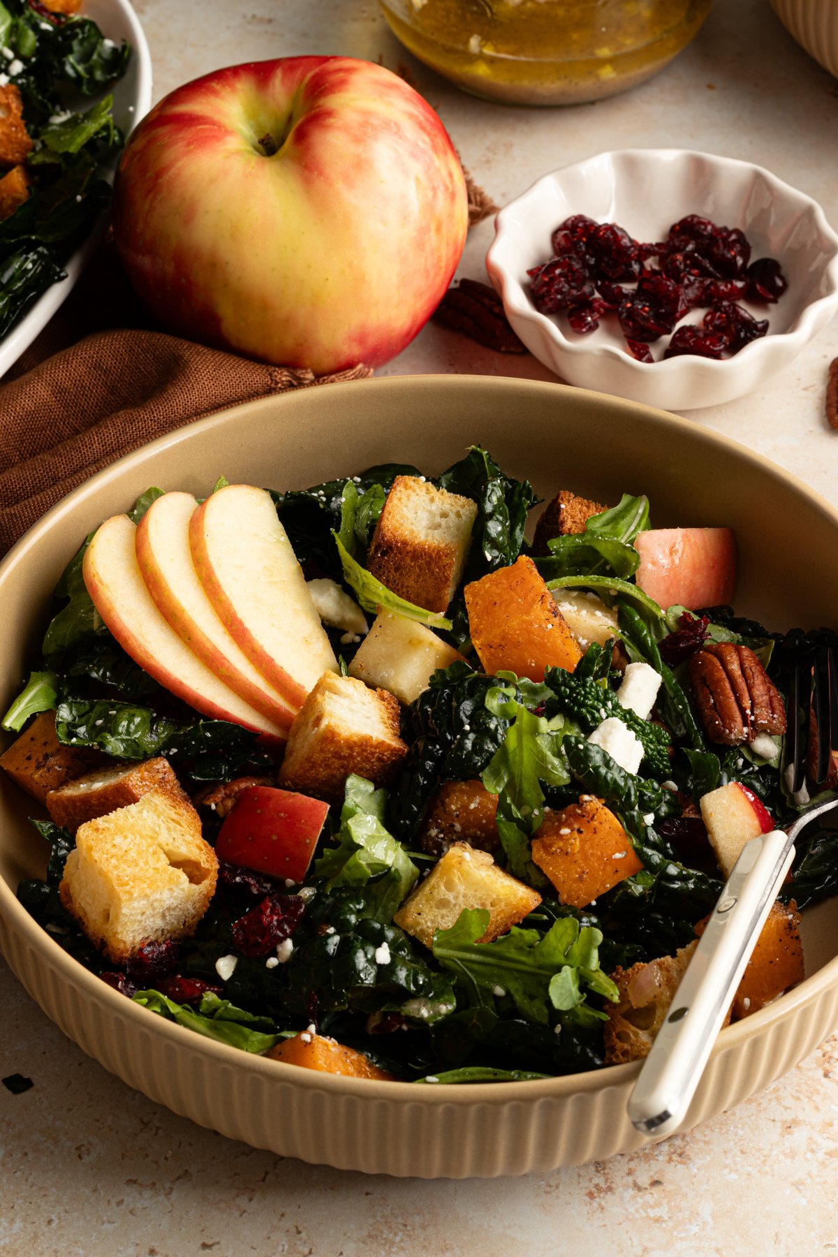 Side view of a single bowl of salad with crisp apple slices, roasted butternut squash, kale, arugula, feta, and crunchy croutons, with an apple and small bowl of dried cranberries in the background.