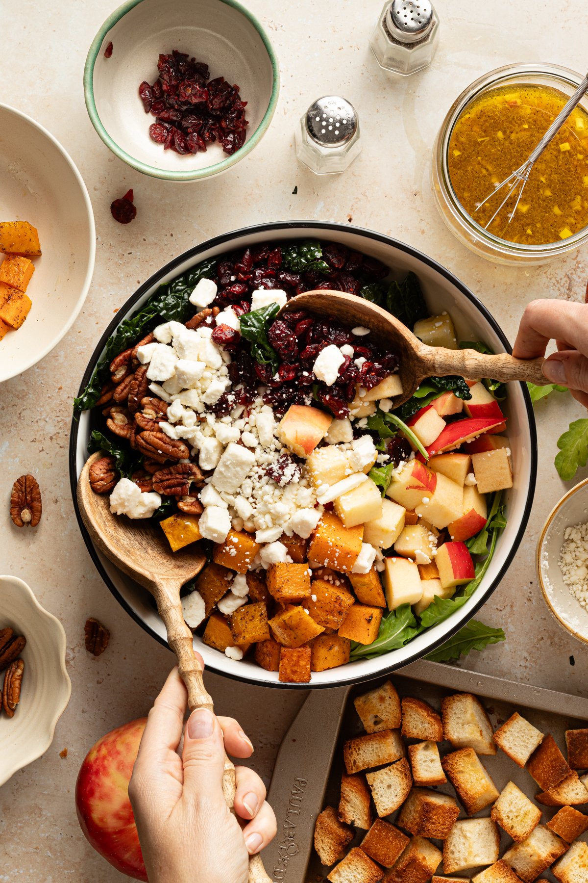 Hands tossing salad with wooden spoons in a large bowl filled with kale, apples, roasted squash, feta, pecans, and cranberries, surrounded by ingredients and toasted bread cubes.