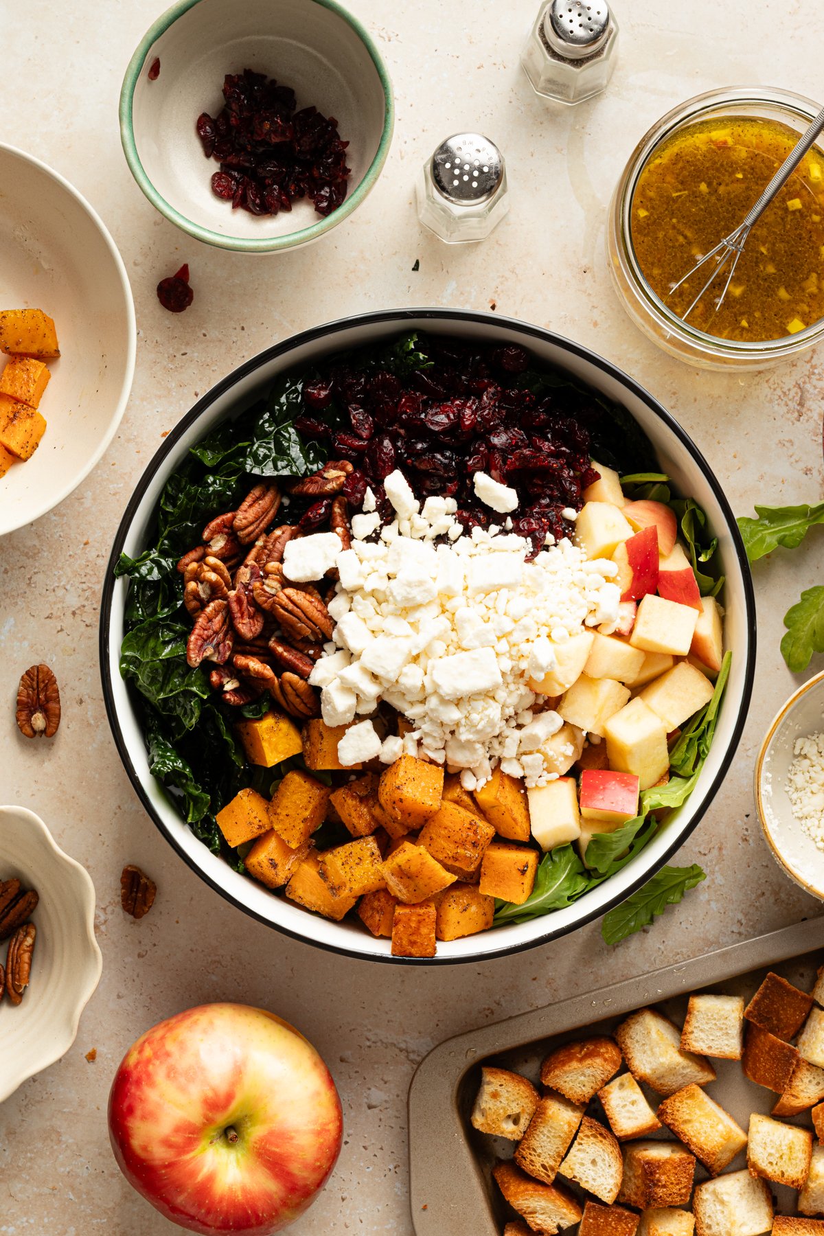 Overhead view of a large salad bowl filled with kale, roasted butternut squash, apples, feta, pecans, and dried cranberries, surrounded by a jar of vinaigrette, toasted bread cubes, and scattered ingredients on a light countertop.