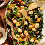 Salad arranged on an oval platter with kale, arugula, roasted butternut squash, apples, pecans, feta, and golden croutons, topped with apple slices and served with wooden salad spoons.