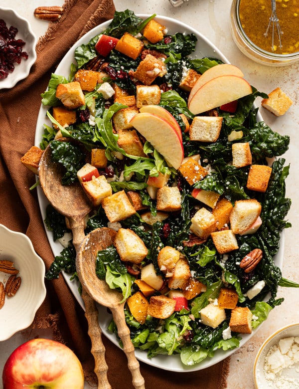 Salad arranged on an oval platter with kale, arugula, roasted butternut squash, apples, pecans, feta, and golden croutons, topped with apple slices and served with wooden salad spoons.
