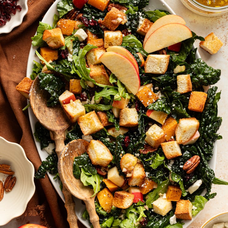Salad arranged on an oval platter with kale, arugula, roasted butternut squash, apples, pecans, feta, and golden croutons, topped with apple slices and served with wooden salad spoons.