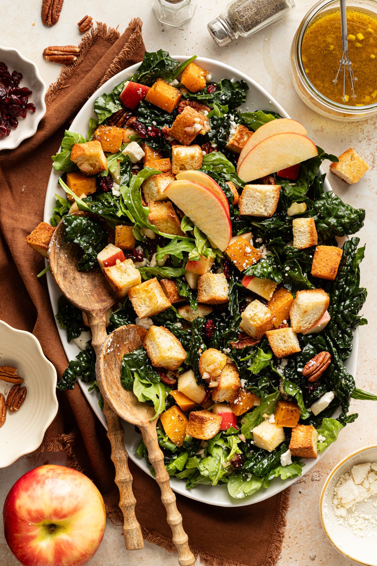 Salad arranged on an oval platter with kale, arugula, roasted butternut squash, apples, pecans, feta, and golden croutons, topped with apple slices and served with wooden salad spoons.