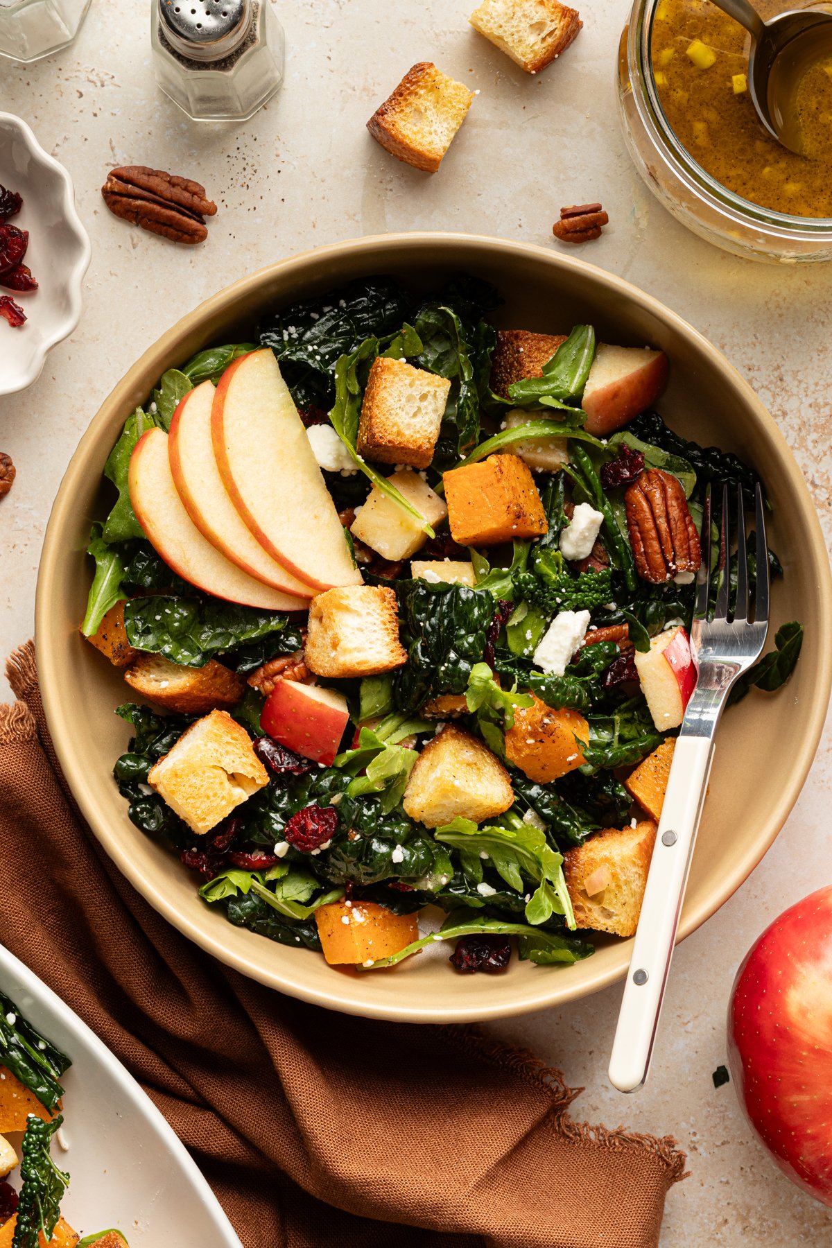 Individual serving of salad in a tan bowl with a fork, featuring kale, roasted butternut squash, croutons, pecans, cranberries, feta, and sliced apples, surrounded by extra dressing and salad ingredients.