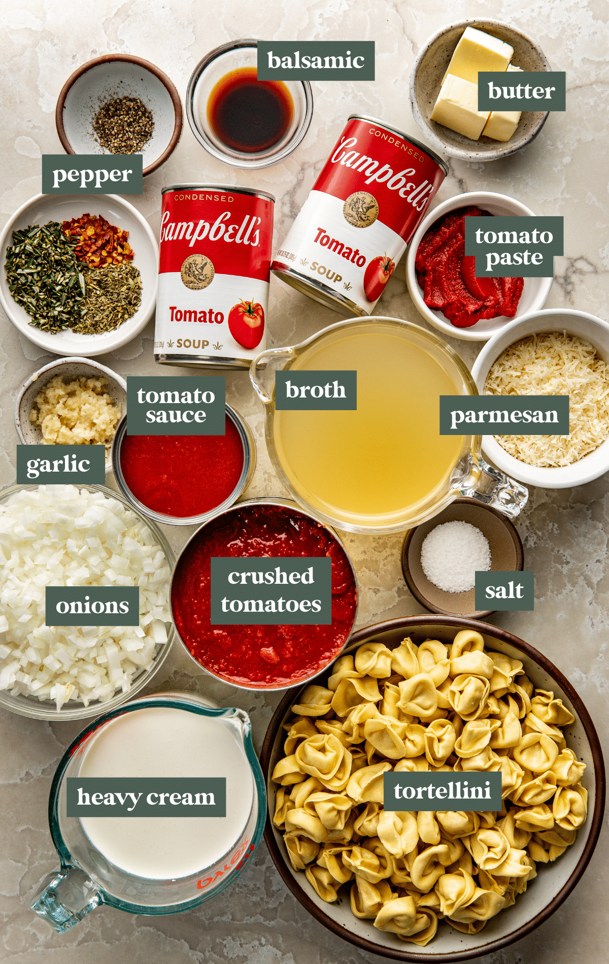 Flat lay of measured soup ingredients including tortellini, canned tomato soup, crushed tomatoes, broth, cream, spices, butter, and chopped onion on a marble surface.