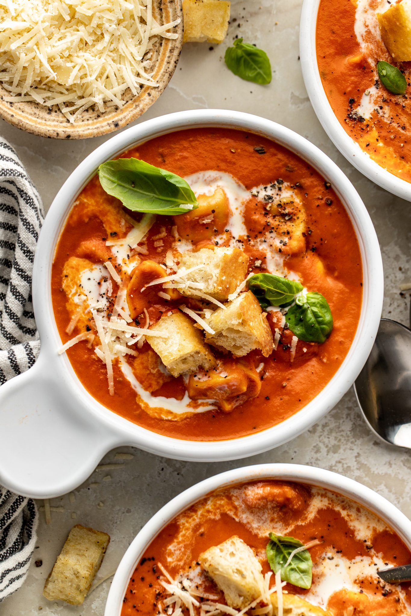 Close-up overhead of tomato tortellini soup in white bowls, garnished with basil, croutons, grated parmesan, and a swirl of cream, next to a small bowl of shredded cheese and striped napkin.
