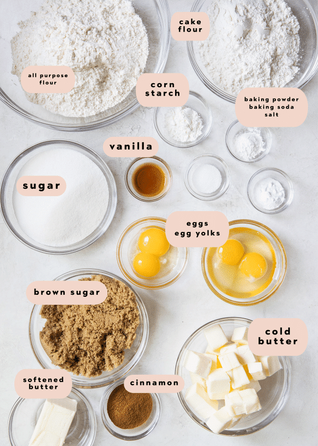 Multiple clear bowls filled with ingredients including flour, cake flour, sugar, corn starch, vanilla, eggs, brown sugar, cold butter, cinnamon, and softened butter plus labels with the name of each ingredient