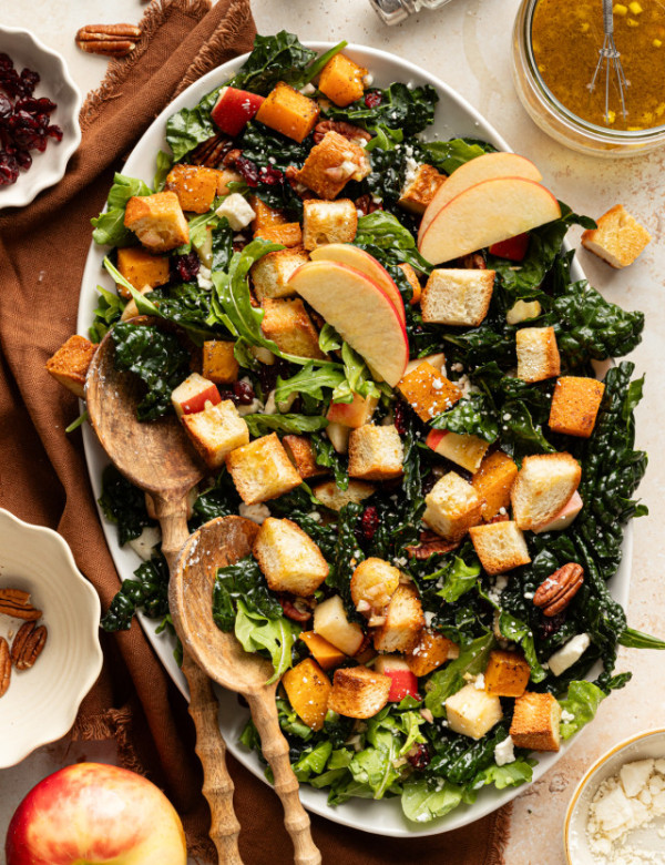 Salad arranged on an oval platter with kale, arugula, roasted butternut squash, apples, pecans, feta, and golden croutons, topped with apple slices and served with wooden salad spoons.