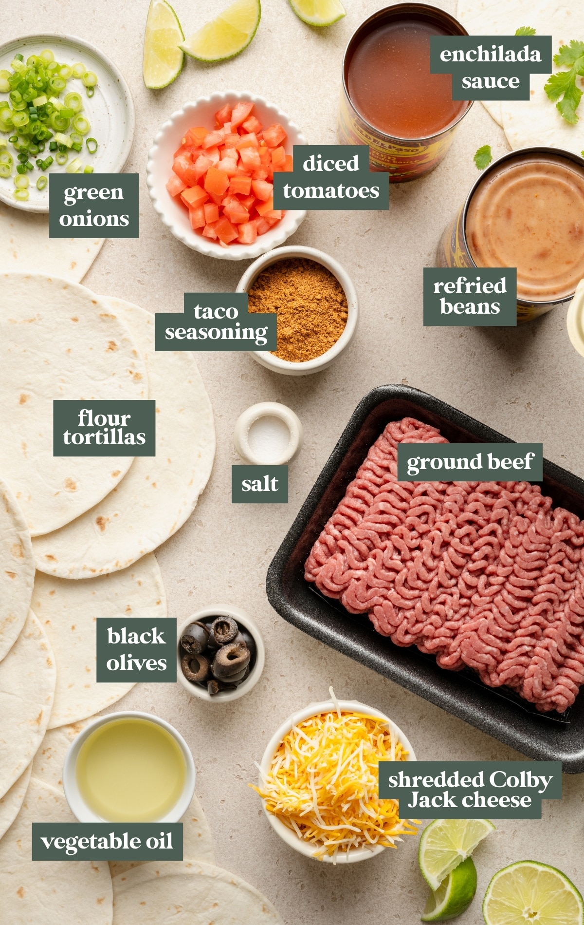 Overhead image showing labeled ingredients for homemade Mexican pizza. Items include ground beef, flour tortillas, refried beans, shredded Colby Jack cheese, enchilada sauce, diced tomatoes, green onions, black olives, taco seasoning, vegetable oil, and salt. Each ingredient is neatly arranged on a light countertop with text labels for easy identification.