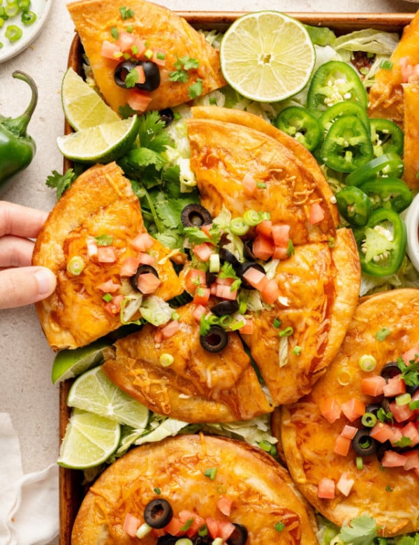 Hand reaching in to grab a slice of Mexican pizza from the tray. The pizzas are fully loaded with melted cheese and fresh toppings like tomatoes, olives, and scallions, and nestled on a bed of lettuce with lime wedges nearby.