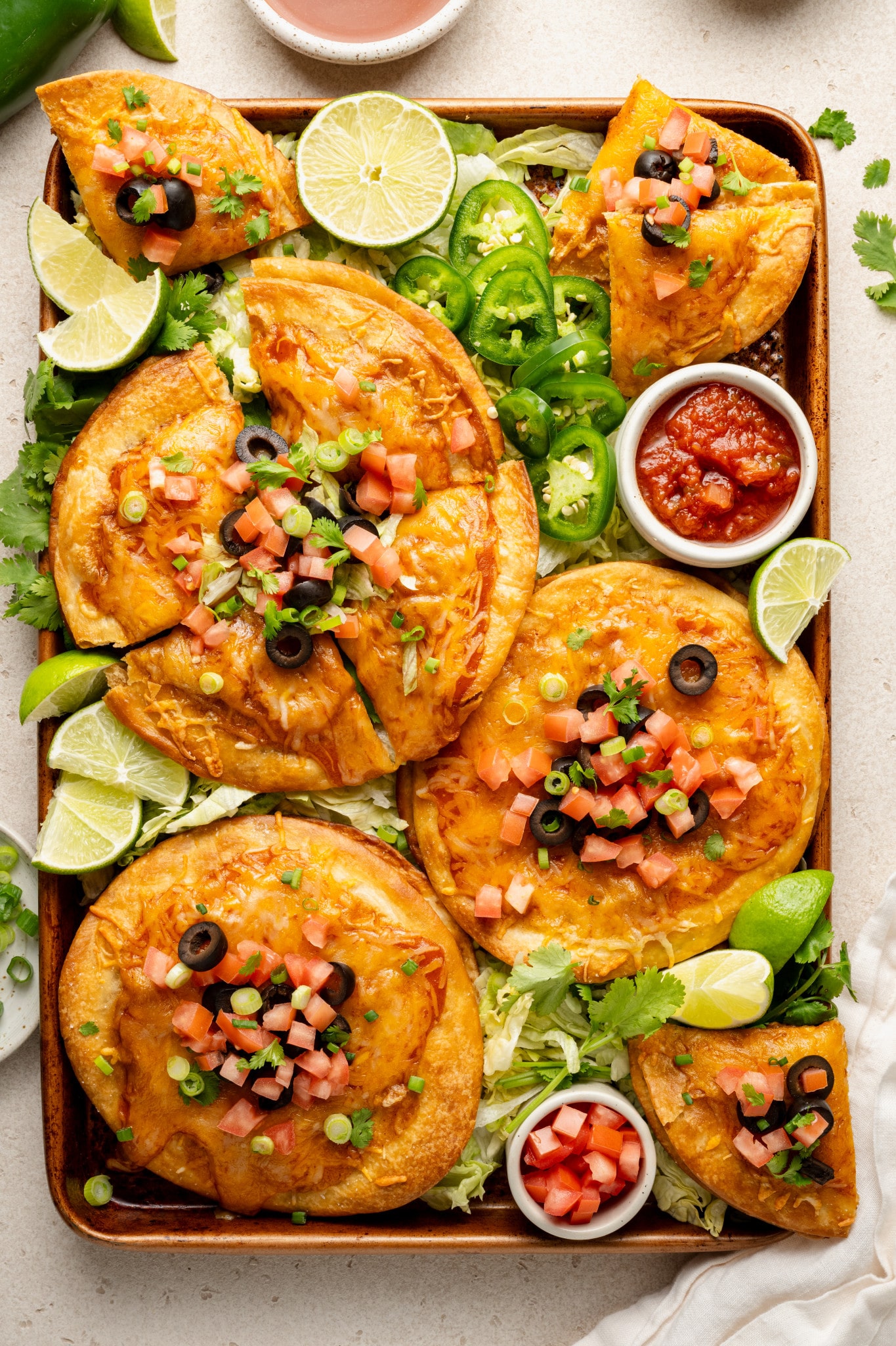 Overhead view of a large tray filled with homemade Mexican pizzas, each topped with melted cheese, diced tomatoes, sliced olives, green onions, and cilantro. The tray is garnished with shredded lettuce, jalapeño slices, lime wedges, and small bowls of salsa and diced tomatoes.