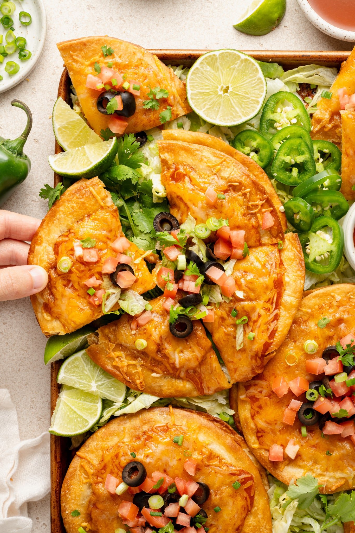 Hand reaching in to grab a slice of Mexican pizza from the tray. The pizzas are fully loaded with melted cheese and fresh toppings like tomatoes, olives, and scallions, and nestled on a bed of lettuce with lime wedges nearby.