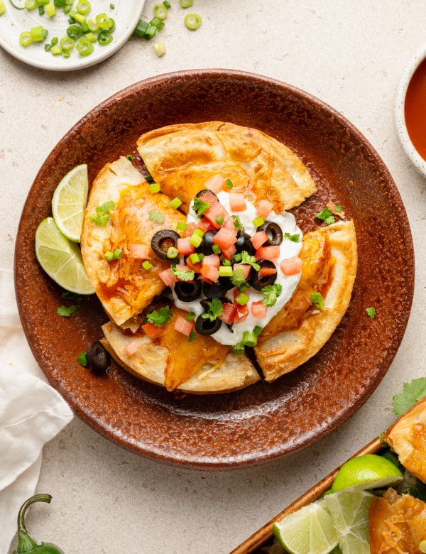 Overhead image of a single serving of Mexican pizza on a rustic brown plate. The pizza is sliced and topped with sour cream, diced tomatoes, black olives, green onions, and cilantro, with lime wedges on the side.