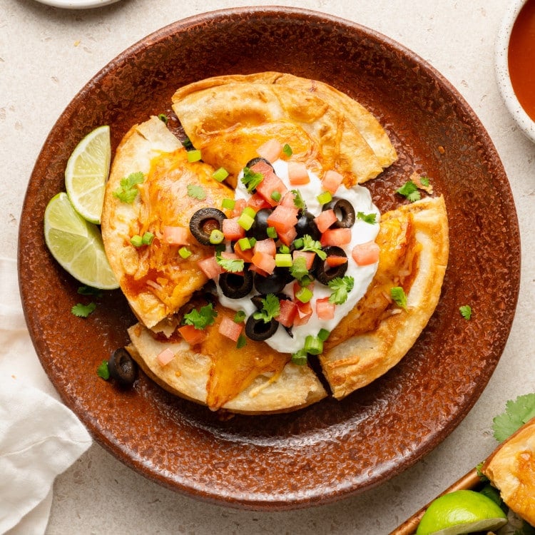 Overhead image of a single serving of Mexican pizza on a rustic brown plate. The pizza is sliced and topped with sour cream, diced tomatoes, black olives, green onions, and cilantro, with lime wedges on the side.
