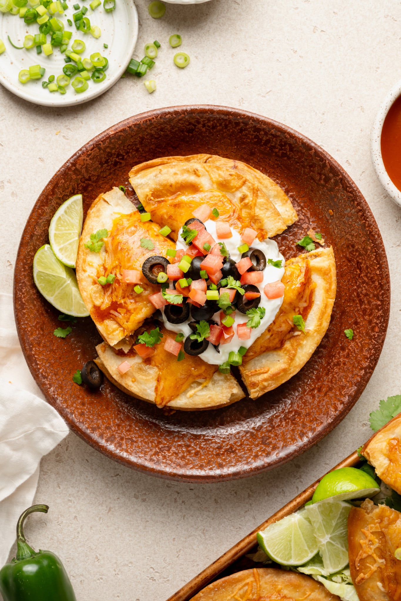 Overhead image of a single serving of Mexican pizza on a rustic brown plate. The pizza is sliced and topped with sour cream, diced tomatoes, black olives, green onions, and cilantro, with lime wedges on the side.