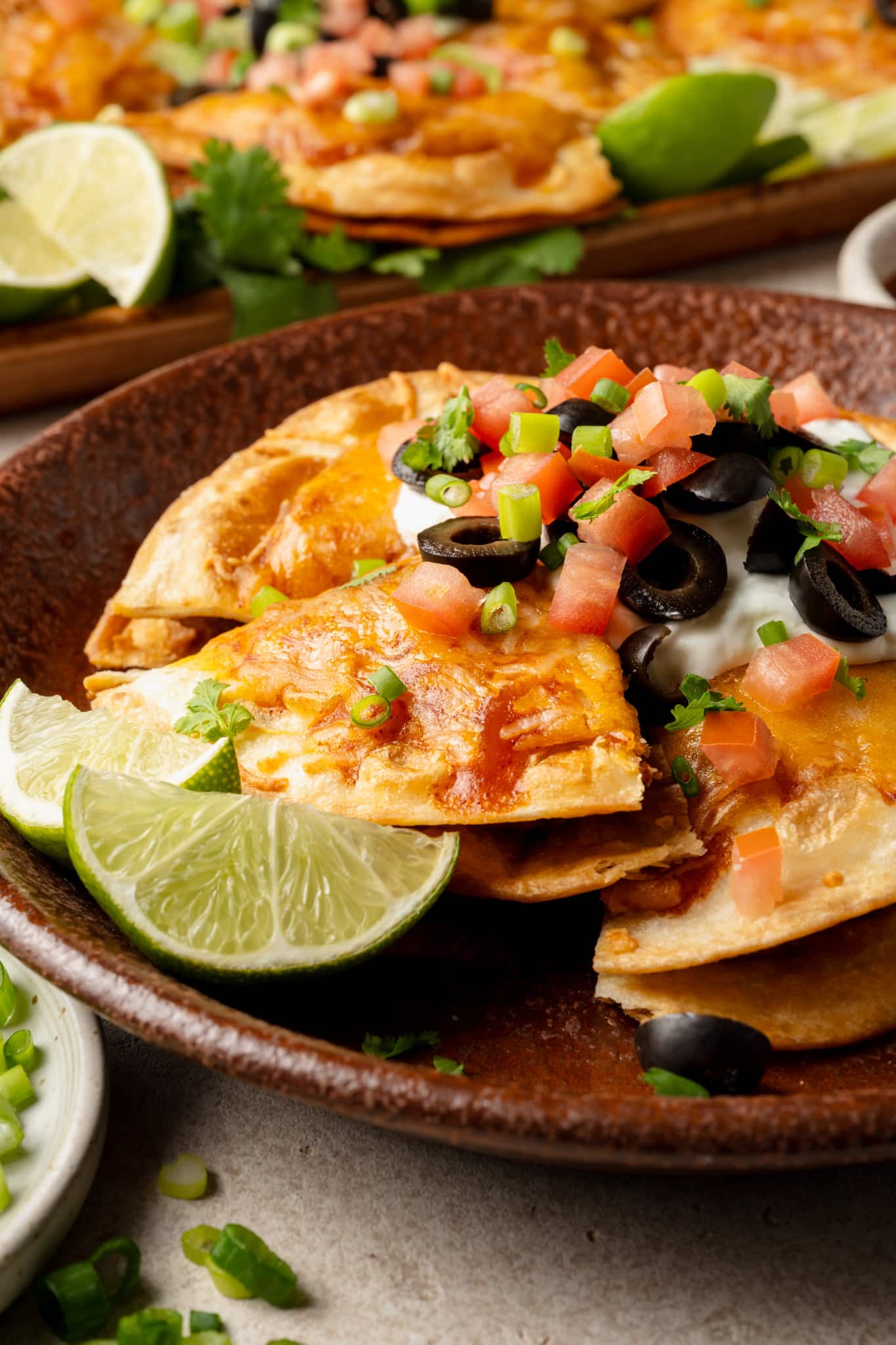Side angle view of a plate of Mexican pizza garnished with lime wedges. The pizza slices are stacked slightly and topped with sour cream, chopped tomatoes, olives, and green onions. Another tray of pizzas is blurred in the background.