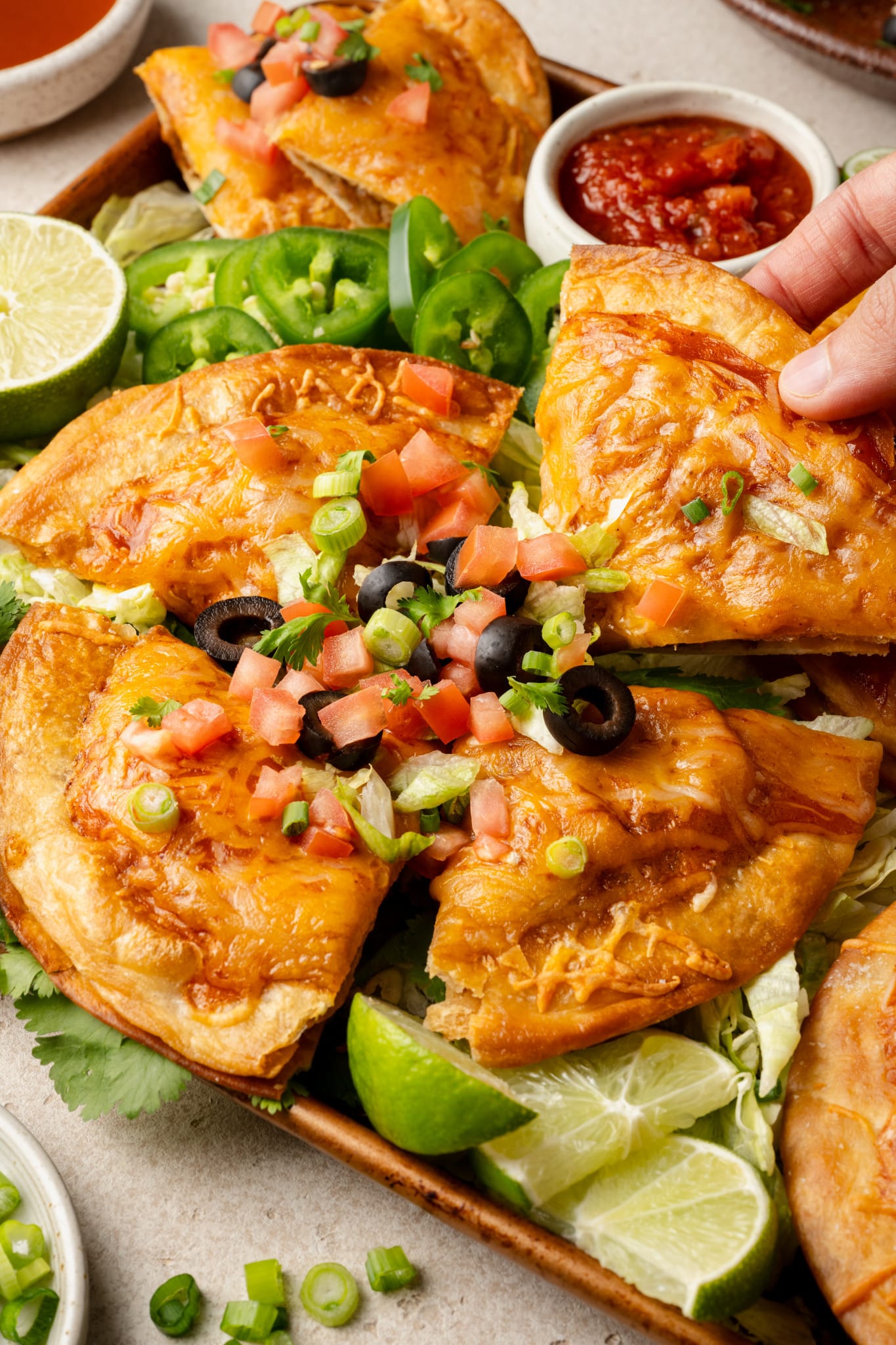 Close-up of a hand lifting a slice of Mexican pizza from the tray, revealing layers of beef and beans inside. The cheesy top is garnished with diced tomatoes, green onions, and olives, surrounded by lime wedges and jalapeños.