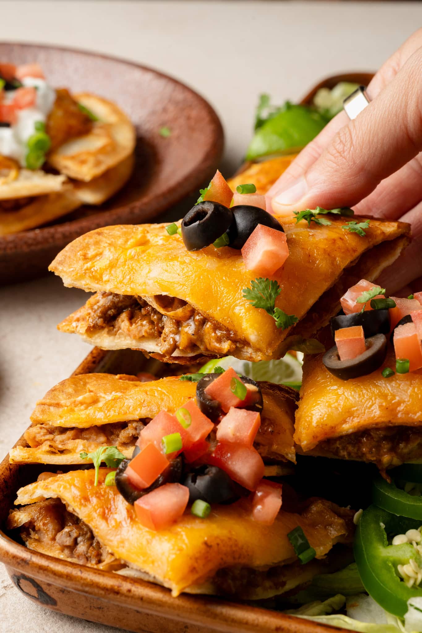 Close-up of a hand lifting a slice of Mexican pizza from a tray. The pizza slice has a crispy golden crust and is layered with seasoned beef and melted cheddar cheese, then topped with diced tomatoes, sliced black olives, green onions, and cilantro.