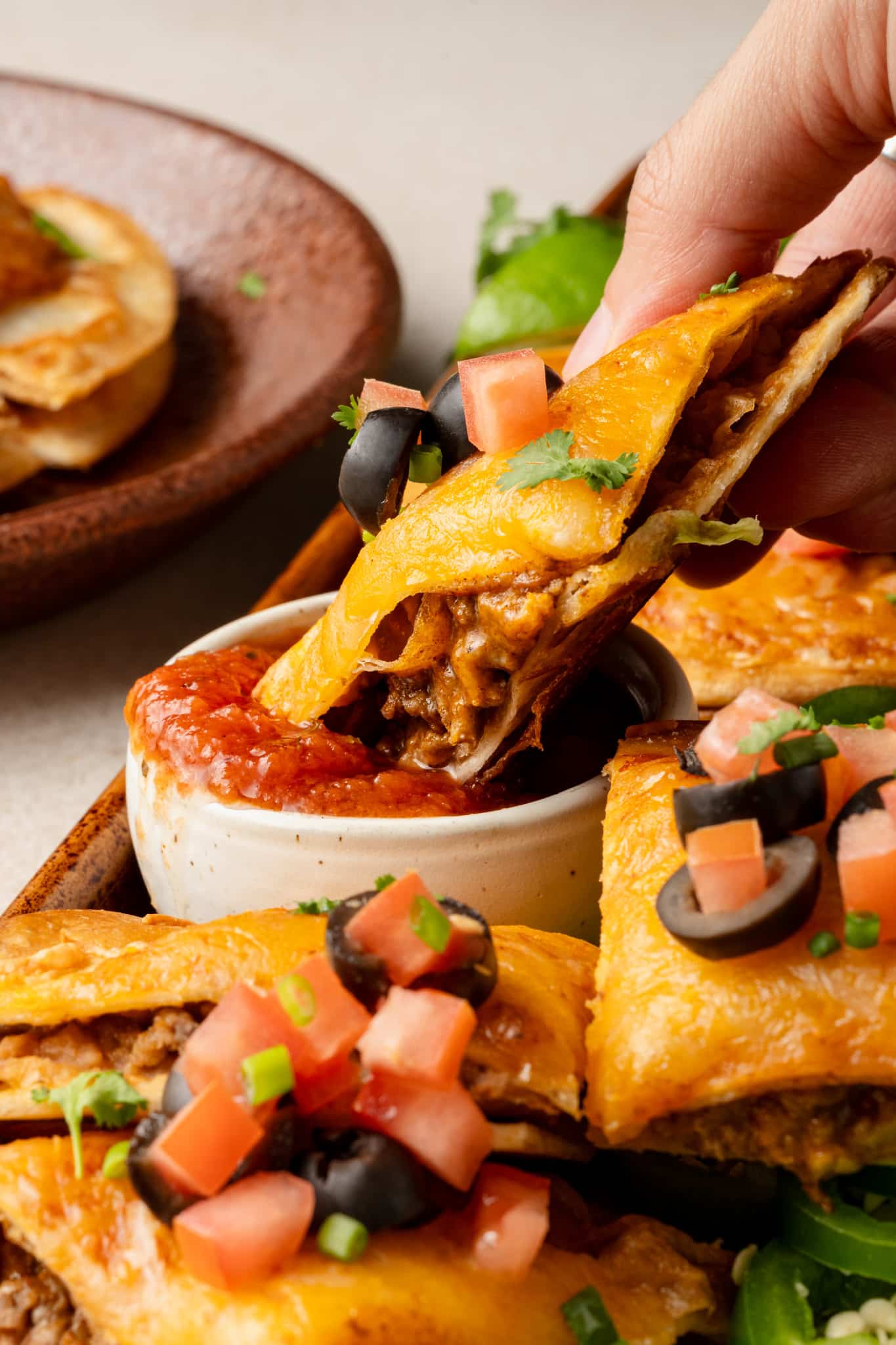 Close-up of a cheesy Mexican pizza slice being dipped into a small bowl of red salsa. The slice is filled with savory beef and topped with cheddar cheese, olives, diced tomatoes, and cilantro, with more pizza slices visible in the background.
