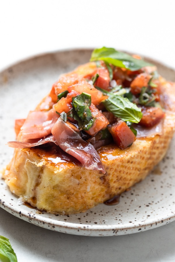 a slice of french toast with Prosciutto, tomatoes and basil on a white speckled plate