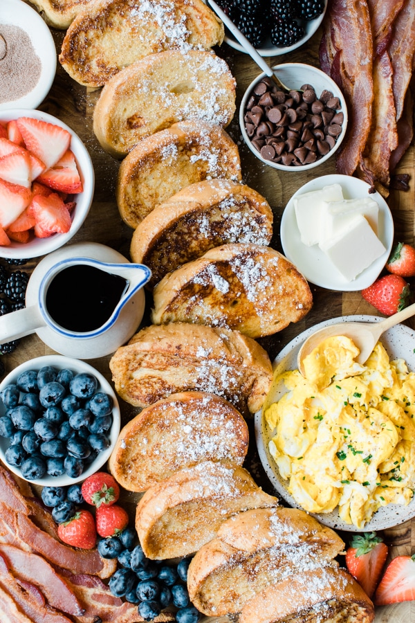 french toast on a cutting board with fresh berries, bacon, scrambled eggs and syrup