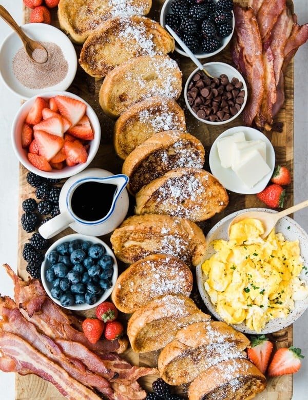a cutting board filled with french toast, fresh berries, bacon and syrup