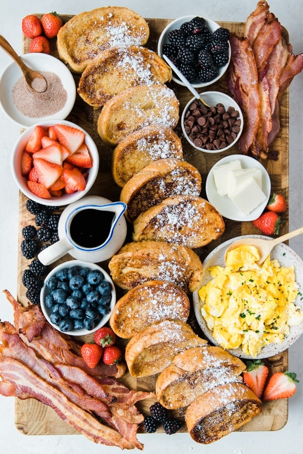 a cutting board filled with french toast, fresh berries, bacon and syrup