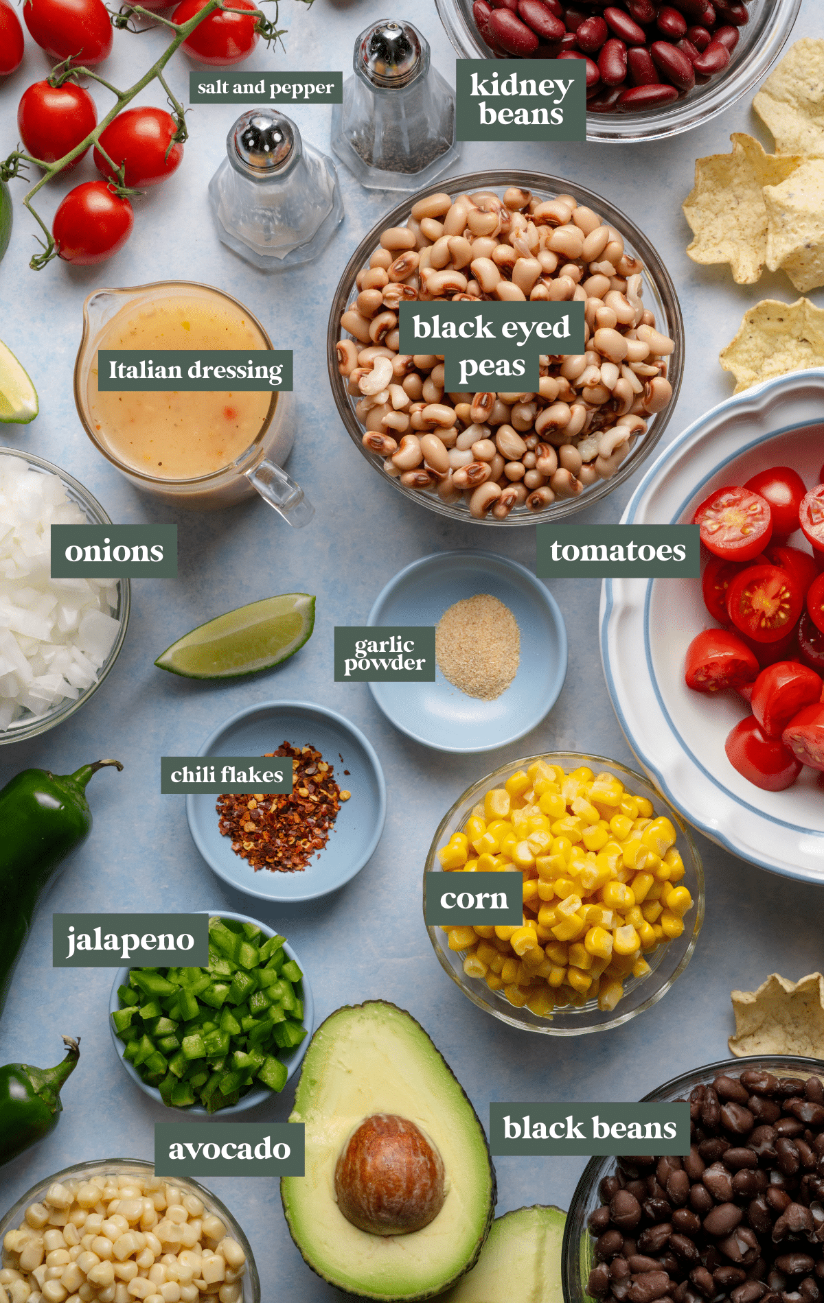 Overhead view of fresh ingredients for cowboy caviar arranged on a blue surface, including beans, corn, cherry tomatoes, onion, avocado, jalapeño, and a small pitcher of dressing.