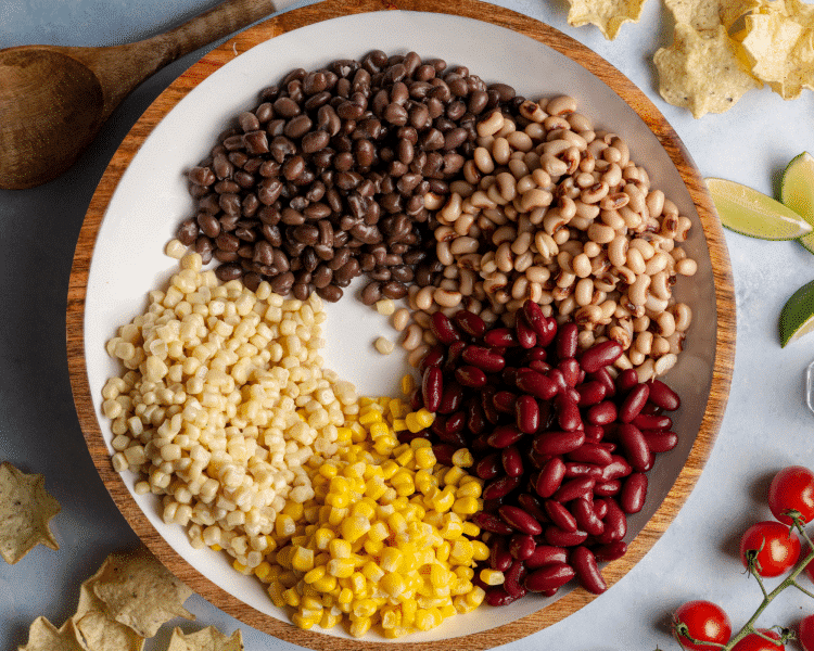 Overhead view of a large white bowl filled with separated piles of black beans, black-eyed peas, red kidney beans, white corn, and yellow corn, ready to be mixed for cowboy caviar.