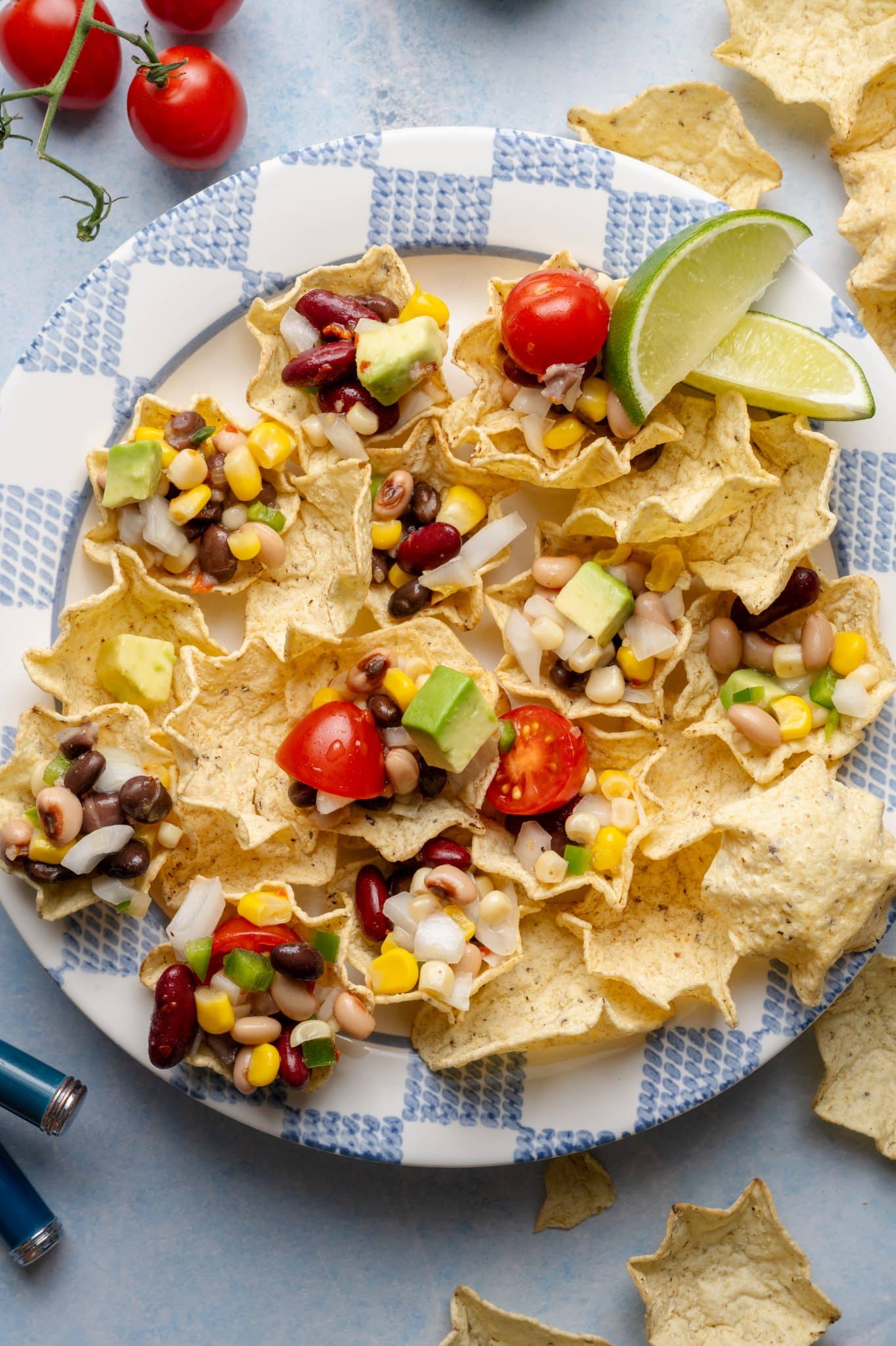 A checkered plate filled with tortilla scoop chips topped with cowboy caviar, garnished with cherry tomatoes, avocado, and lime wedges.
