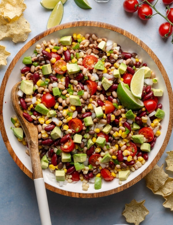 Mixed cowboy caviar in a large bowl with a wooden spoon, showing vibrant beans, corn, tomatoes, jalapeños, and diced avocado.