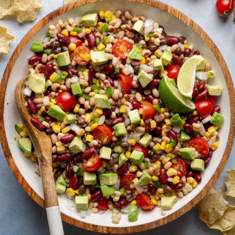 Mixed cowboy caviar in a large bowl with a wooden spoon, showing vibrant beans, corn, tomatoes, jalapeños, and diced avocado.
