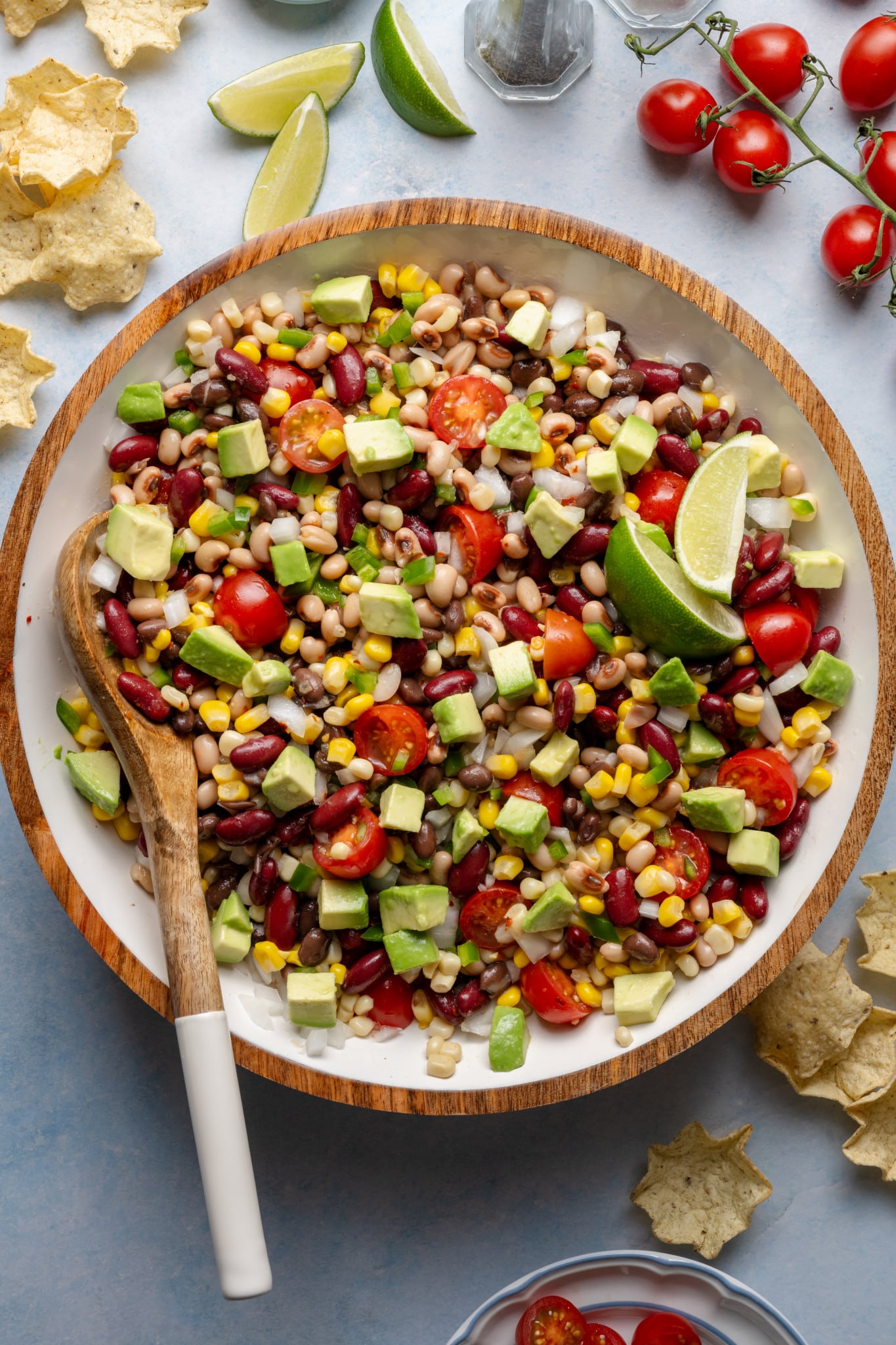 Mixed cowboy caviar in a large bowl with a wooden spoon, showing vibrant beans, corn, tomatoes, jalapeños, and diced avocado.