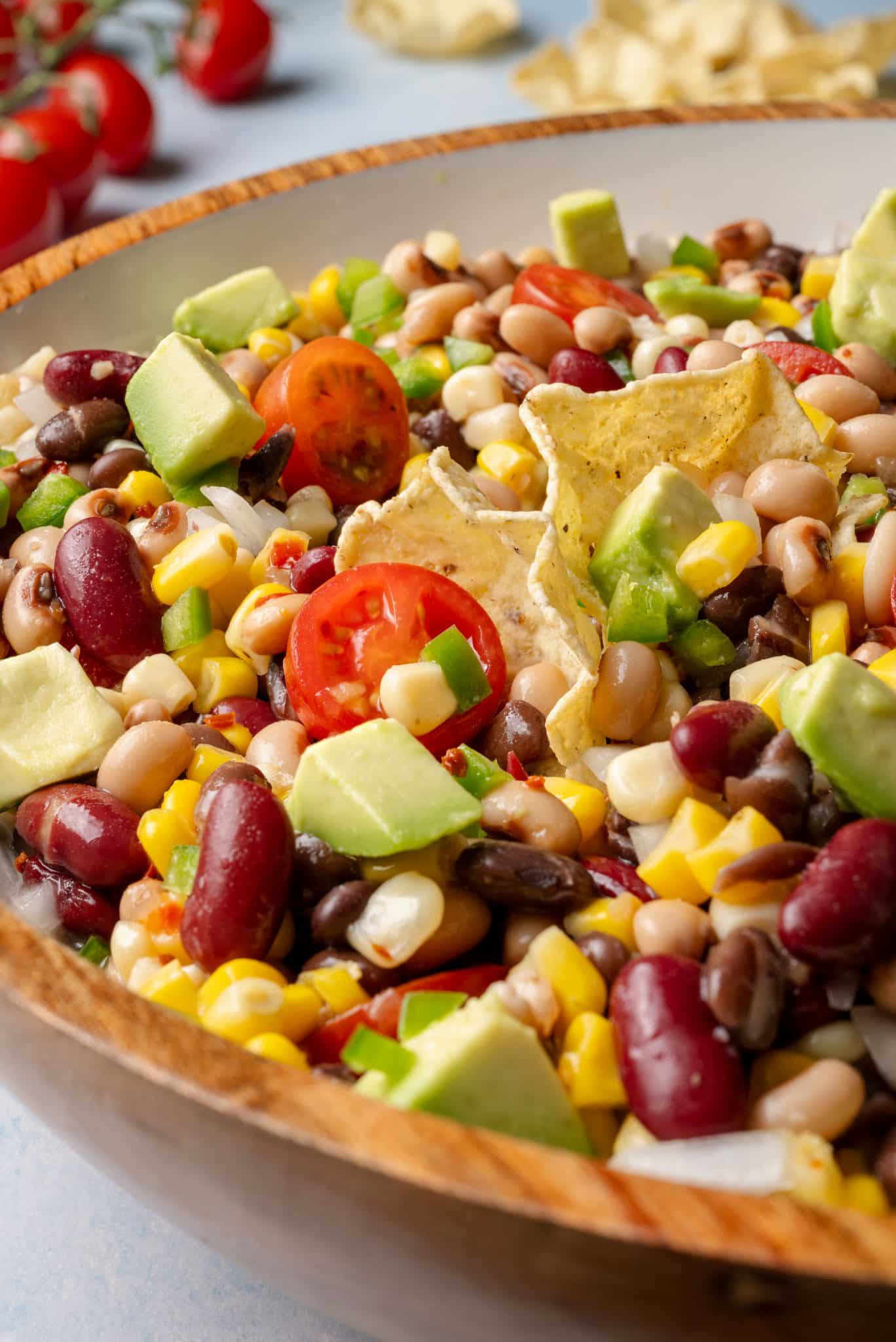 Close-up of cowboy caviar in a wooden bowl with tortilla chips and a wooden spoon, showcasing fresh veggies, beans, corn, and avocado.