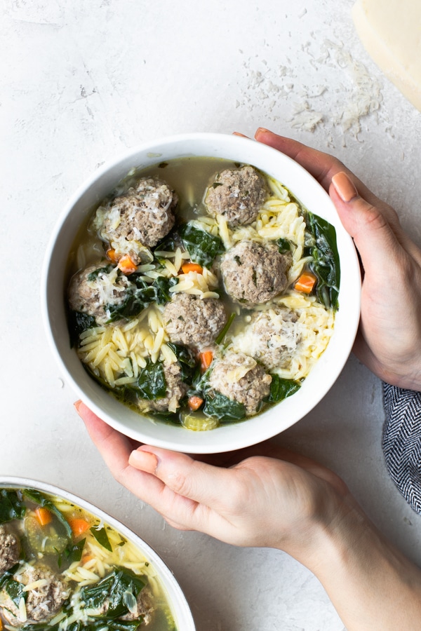 A woman's hands holding a white bowl filled with Italian wedding soup 